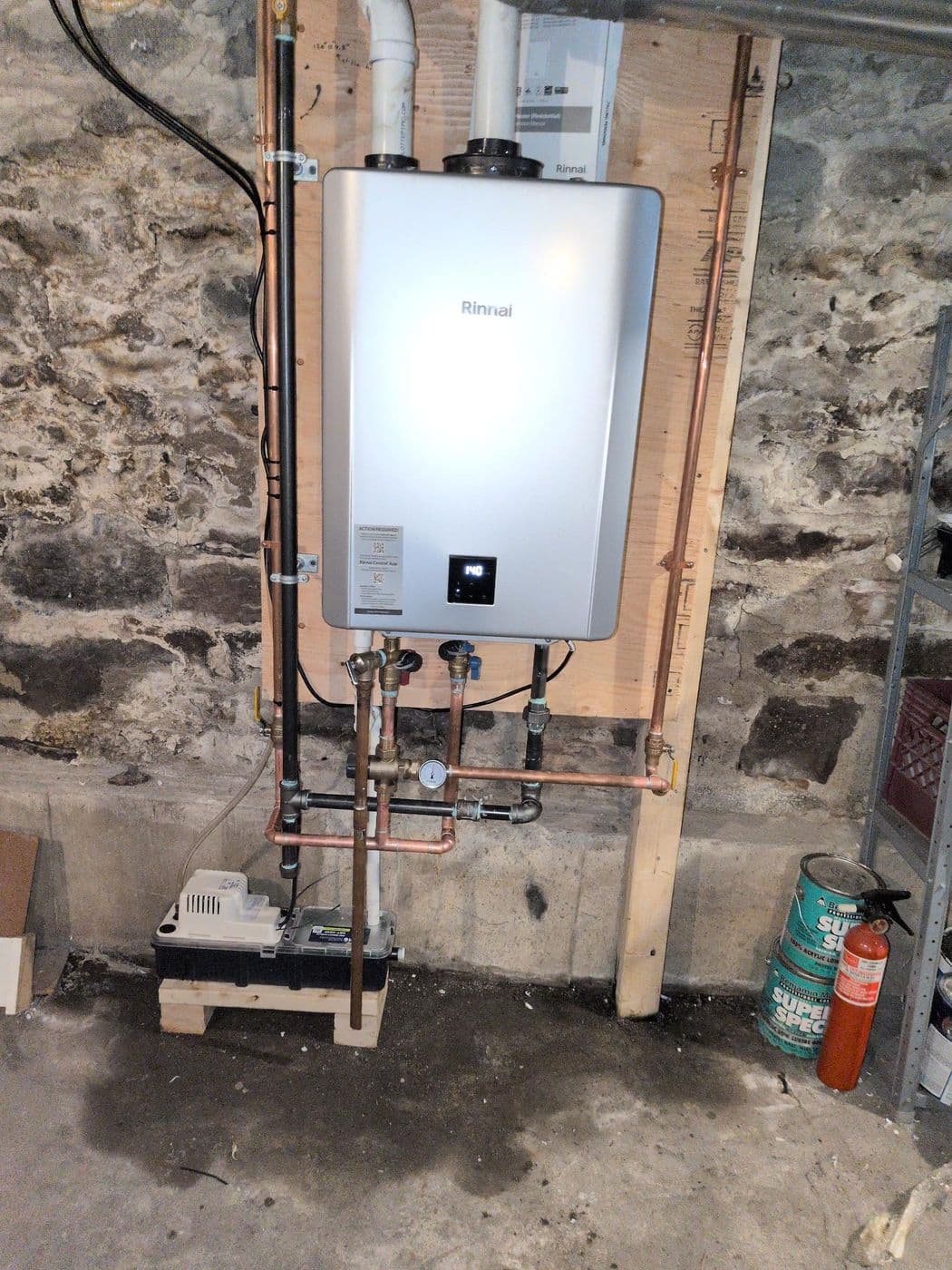 Tankless water heater mounted on a wooden panel against an old stone foundation wall